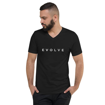 A man with a beard has his hands in his pockets while wearing an Evolve Unisex Short Sleeve V-Neck T-Shirt from Evolve Key West. The shirt is crafted from premium material and features the word "EVOLVE" printed in white across the chest, highlighting its ethical sourcing. He glances down to his right.