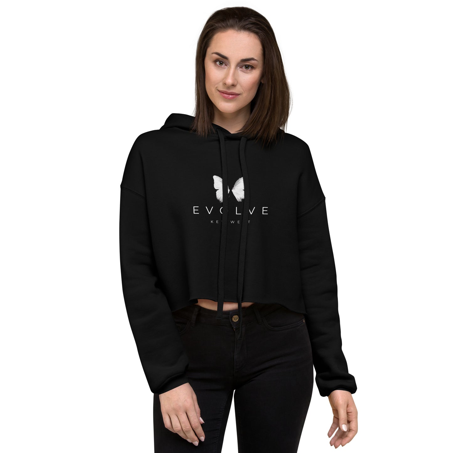 A person with shoulder-length brown hair is wearing a black Evolve Key West Crop Hoodie, featuring "EVOLVE" and a butterfly design on the front, showcasing their commitment to sustainable fashion. With drawstrings adding to the hoodie’s details, they stand against a plain white background, one hand casually tucked in their pocket.